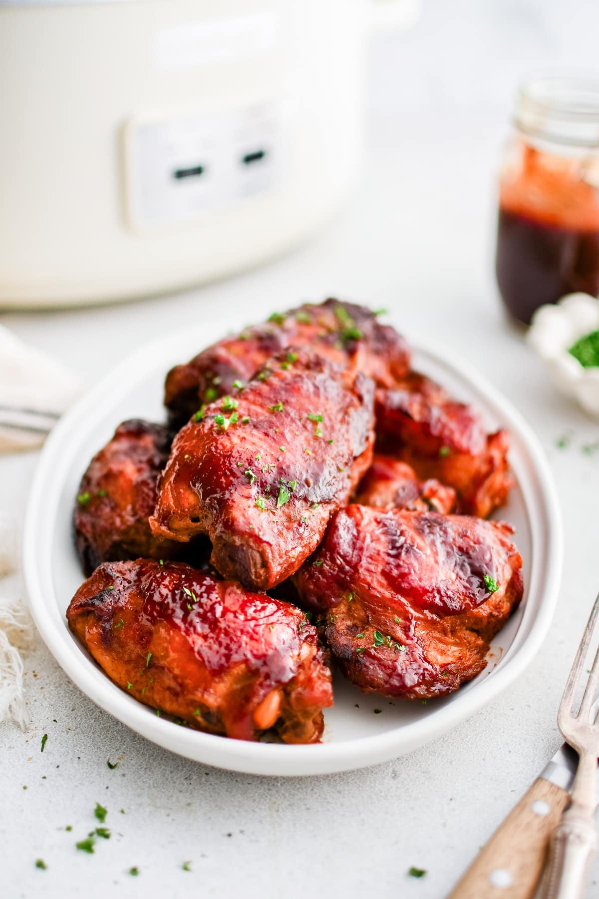 A plate of slow cooker BBQ chicken thighs garnished with fresh parsley. A small bowl of extra BBQ sauce is nearby for dipping.
