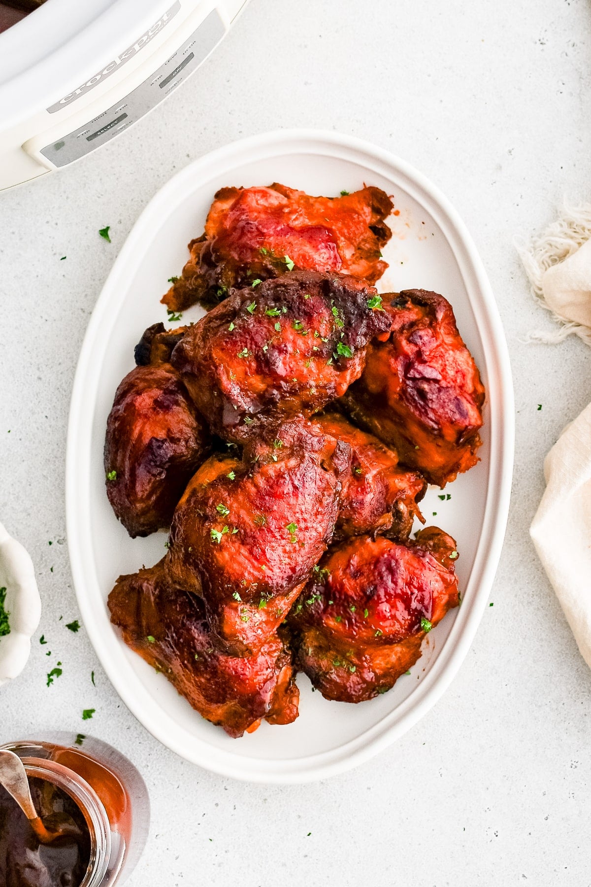 A plate of slow cooker BBQ chicken thighs garnished with fresh parsley. A small bowl of extra BBQ sauce is nearby for dipping.