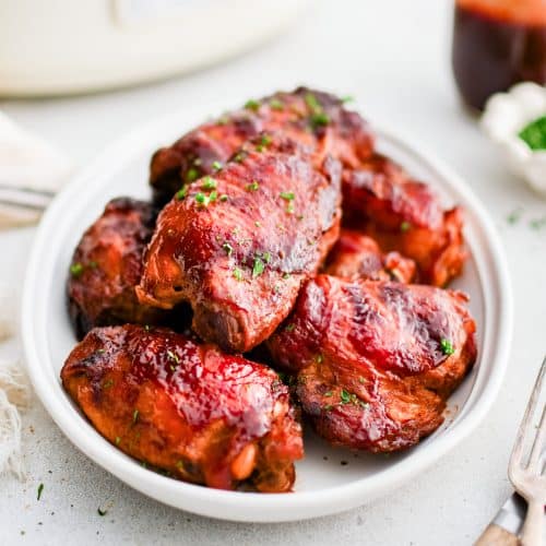 A plate of slow cooker BBQ chicken thighs garnished with fresh parsley. A small bowl of extra BBQ sauce is nearby for dipping.