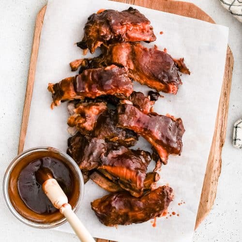 Slow-cooked baby back pork ribs on a large cutting board lined with parchment paper coated in sweet and tangy BBQ sauce.