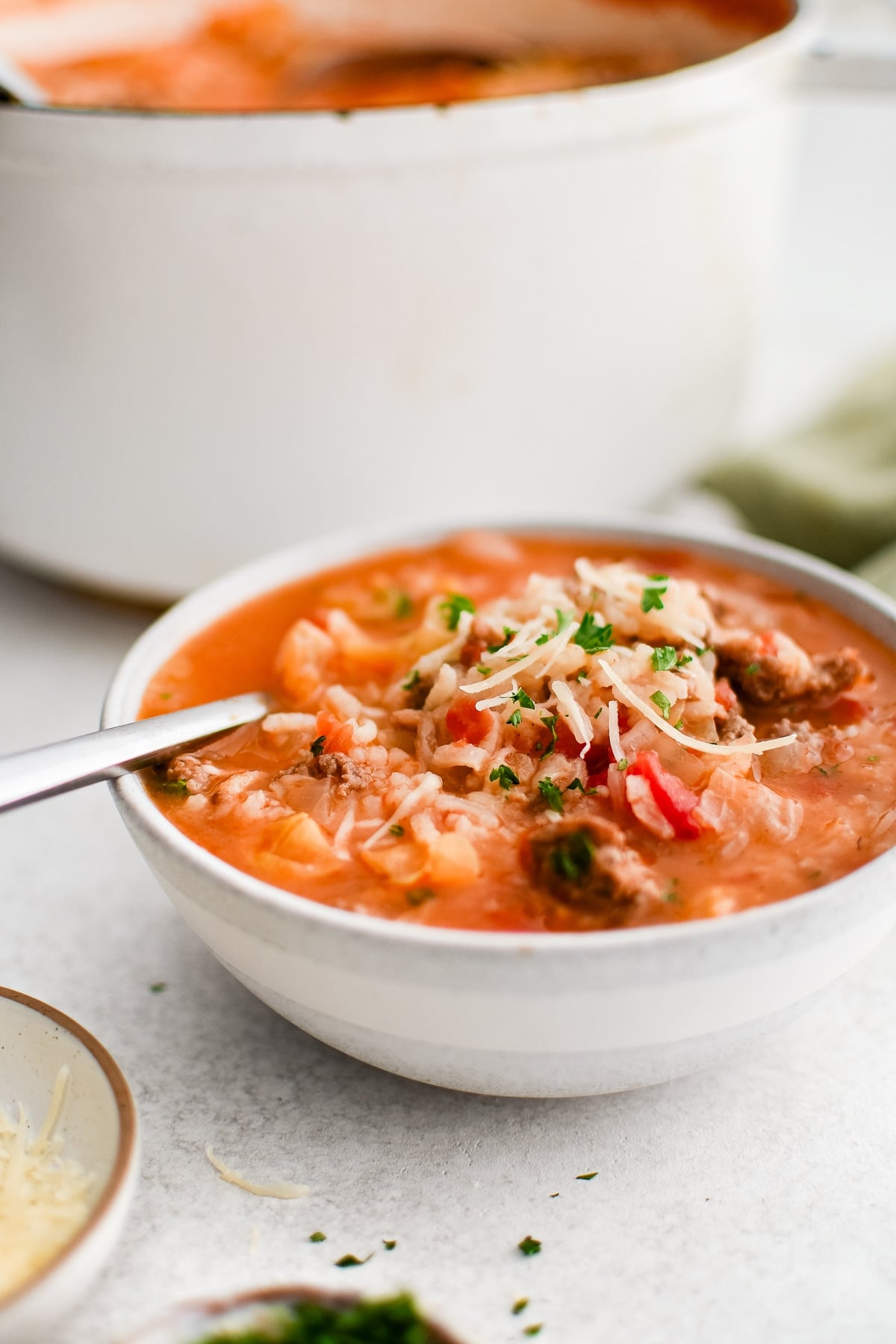 A bowl of Cabbage Roll Soup served hot, with a deep reddish broth surrounding softened cabbage, crumbled ground meat, and fluffy rice. The dish is garnished with freshly grated parmesan cheese and chopped parsley, adding color and texture. A spoon rests inside, ready for eating.