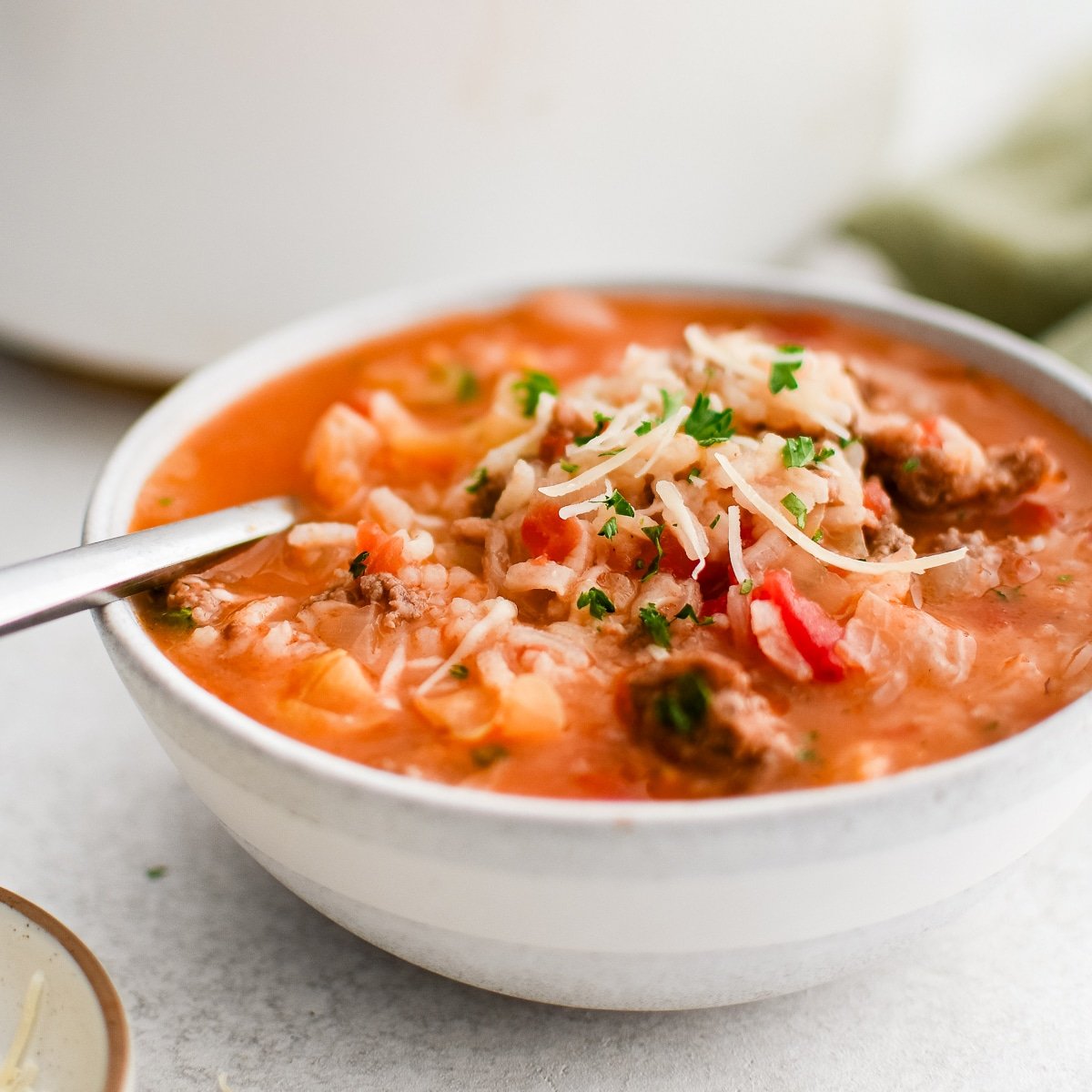 A bowl of Cabbage Roll Soup served hot, with a deep reddish broth surrounding softened cabbage, crumbled ground meat, and fluffy rice. The dish is garnished with freshly grated parmesan cheese and chopped parsley, adding color and texture. A spoon rests inside, ready for eating.