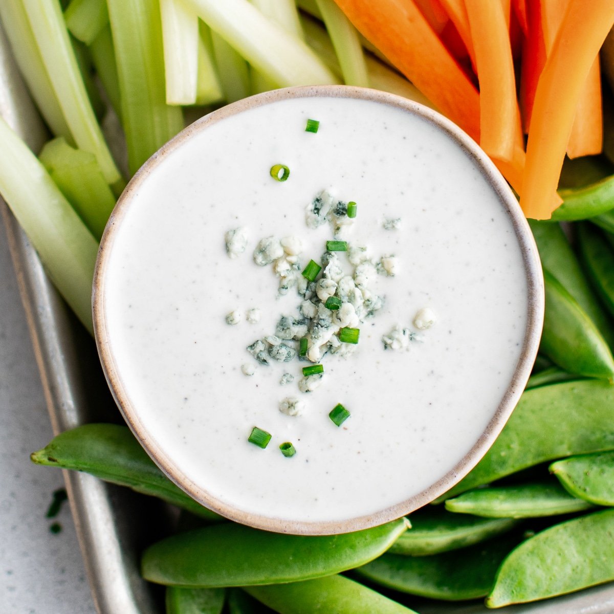A small white bowl filled with creamy blue cheese dressing, sprinkled with blue cheese crumbles and chopped chives, surrounded by fresh raw vegetables like sugar snap peas, carrots, and celery for dipping.