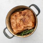 Pork shoulder nestled in a Dutch oven with herbs and braising liquid.