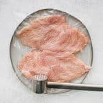 Chicken breasts being pounded under plastic wrap to even thickness on a cutting board.