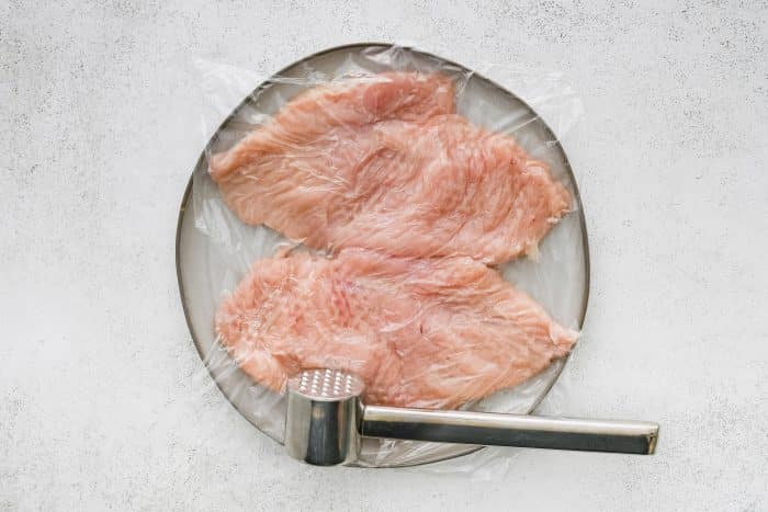 Chicken breasts being pounded under plastic wrap to even thickness on a cutting board.