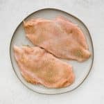 Raw chicken breast cutlets evenly seasoned with spices on a white plate.
