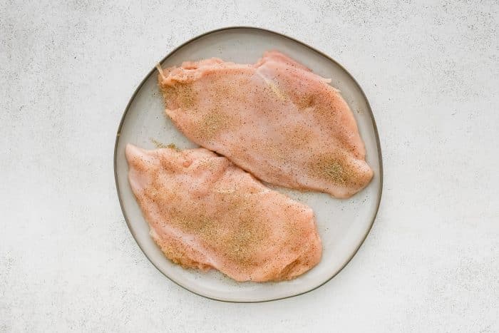 Raw chicken breast cutlets evenly seasoned with spices on a white plate.