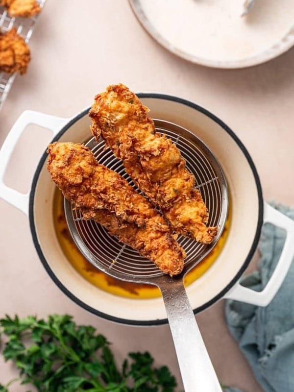 Perfectly golden brown and crispy chicken tenders being lifted from a large pot of sizzling oil in a spider strainer.