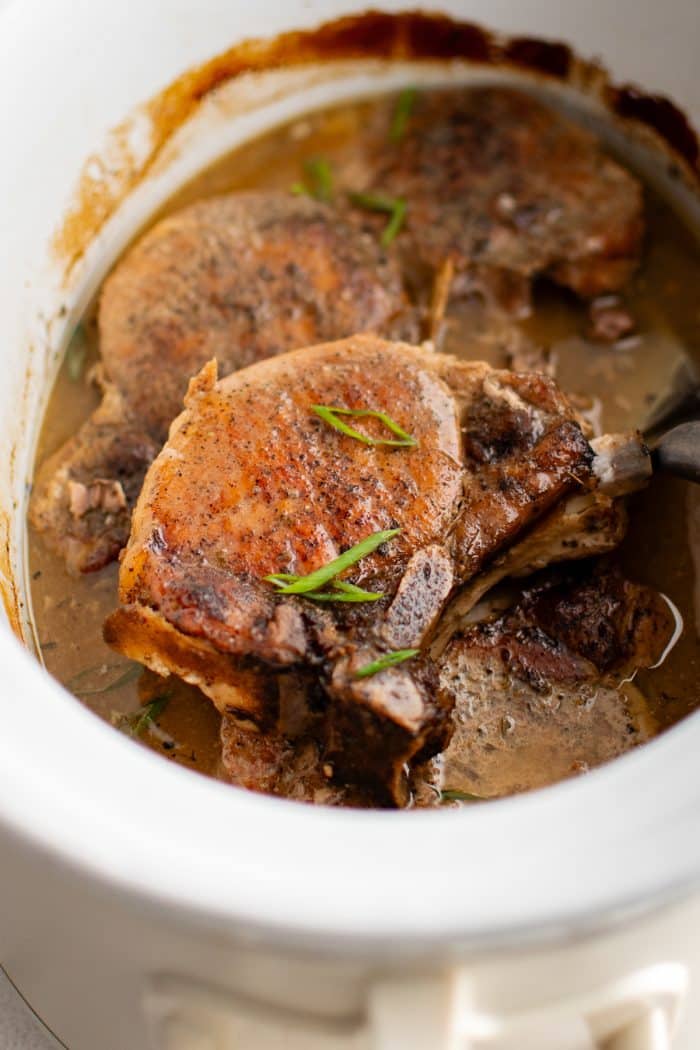 Juicy cooked bone-in rib chops in a slow cooker partially covered in thickened Dijon wine sauce and garnished with sliced green onions.