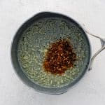 Close-up of the saucepan showing the red pepper flakes and smoked paprika just added to the melted butter and garlic. The mixture is vivid red and gently sizzling, creating a deep, fragrant base for the honey glaze.