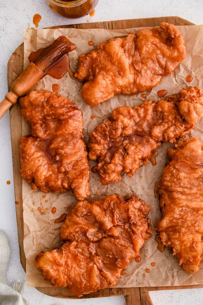 A rustic wooden cutting board piled high with golden-brown fried chicken, each piece generously coated in a glossy hot honey glaze. The sauce glistens under soft lighting, accentuating the crispy texture of the crust. Small drips of honey pool on the board, and a few sprigs of fresh parsley add a pop of color. A small bowl of extra hot honey sits nearby for dipping or drizzling.