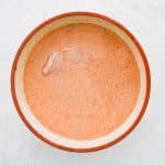 A large glass bowl filled with a pale orange buttermilk mixture speckled with paprika and cayenne. Thin chicken breasts are fully coated and immersed in the marinade.