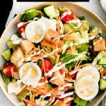 A big, round serving bowl brimming with a colorful house salad, perfectly layered with fresh lettuce, crisp cucumbers, juicy tomatoes, shredded carrots, Cheddar cheese, sliced hard-boiled eggs, and golden croutons, all finished with a generous drizzle of ranch dressing.