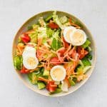 A fresh house salad being assembled step by step, starting with a bed of crisp lettuce and topped with sliced cucumbers, grape tomatoes, shredded carrots, shredded cheddar cheese, hard-boiled eggs, and crunchy croutons.
