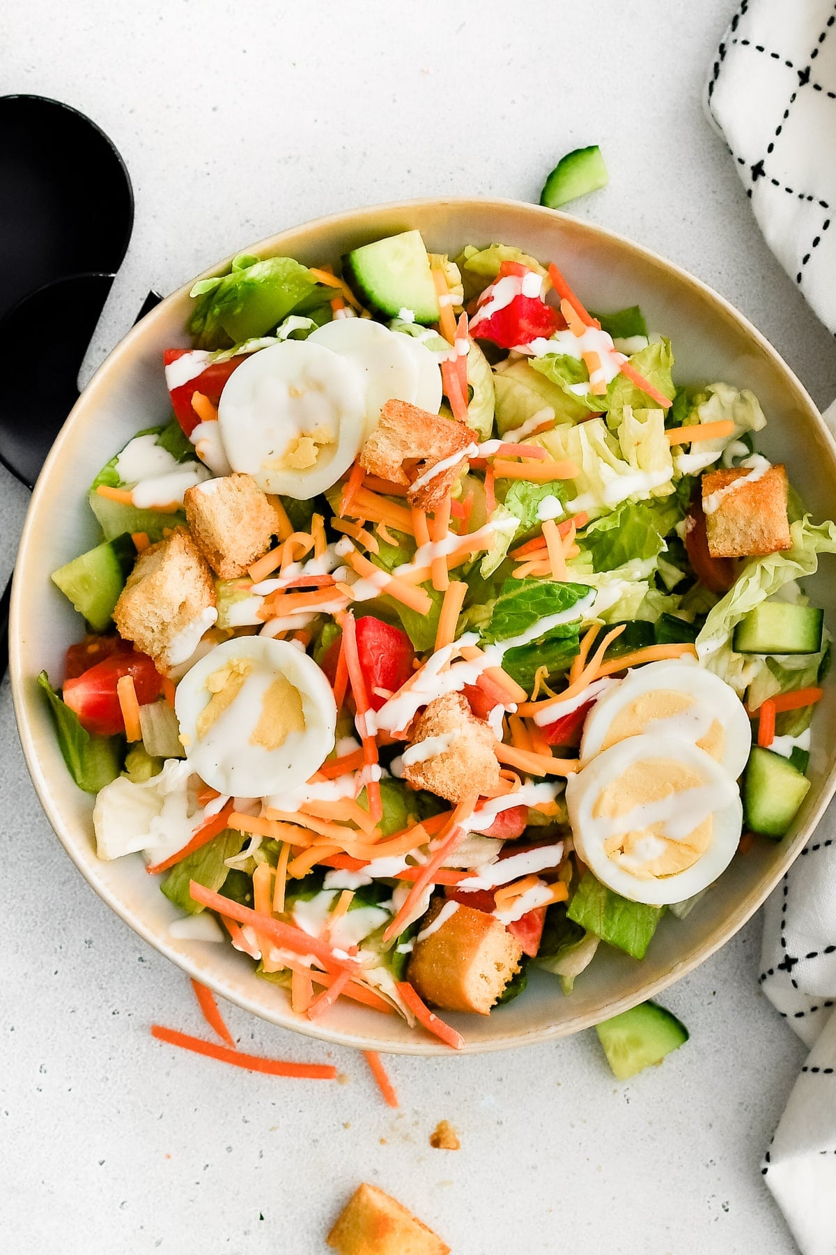 A big, round serving bowl brimming with a colorful house salad, perfectly layered with fresh lettuce, crisp cucumbers, juicy tomatoes, shredded carrots, Cheddar cheese, sliced hard-boiled eggs, and golden croutons, all finished with a generous drizzle of ranch dressing.