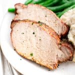 A detailed close-up of two thick slices of pork loin, showing the juicy interior and seasoned crust. The slices rest beside smooth mashed potatoes and vibrant green beans, all on a white plate.