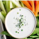 A small white bowl filled with creamy blue cheese dressing, sprinkled with blue cheese crumbles and chopped chives, surrounded by fresh raw vegetables like sugar snap peas, carrots, and celery for dipping.
