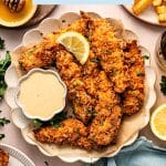 A full serving of golden, crispy buttermilk chicken tenders presented on a round plate with a small bowl of honey mustard dipping sauce, a side plate of French fries, fresh lemon slices, and a soft blue cloth napkin. Additional plates of chicken tenders surround the main dish, making this a perfect meal for sharing or game day.