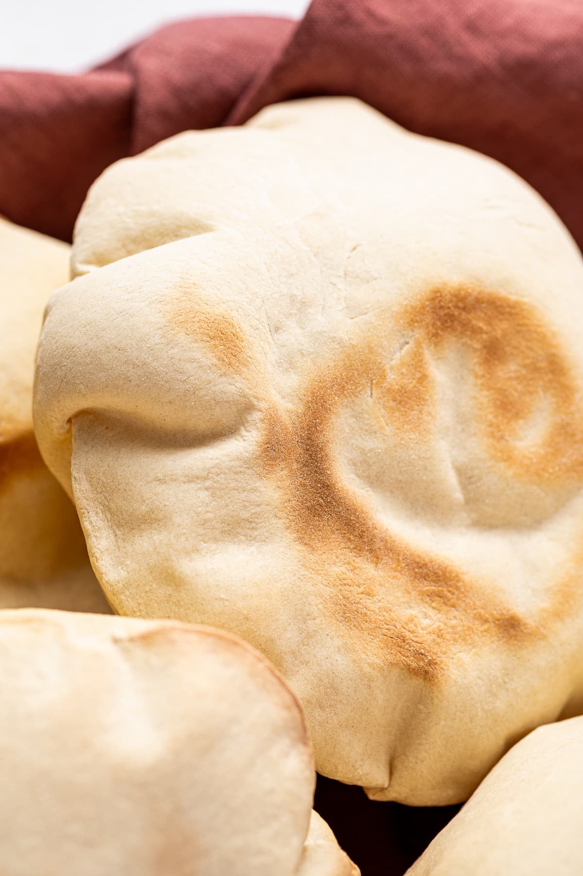 Close-up of a puffed homemade pita bread with golden brown toasted spots, resting against a reddish-brown linen cloth with other pita breads partially visible around it.