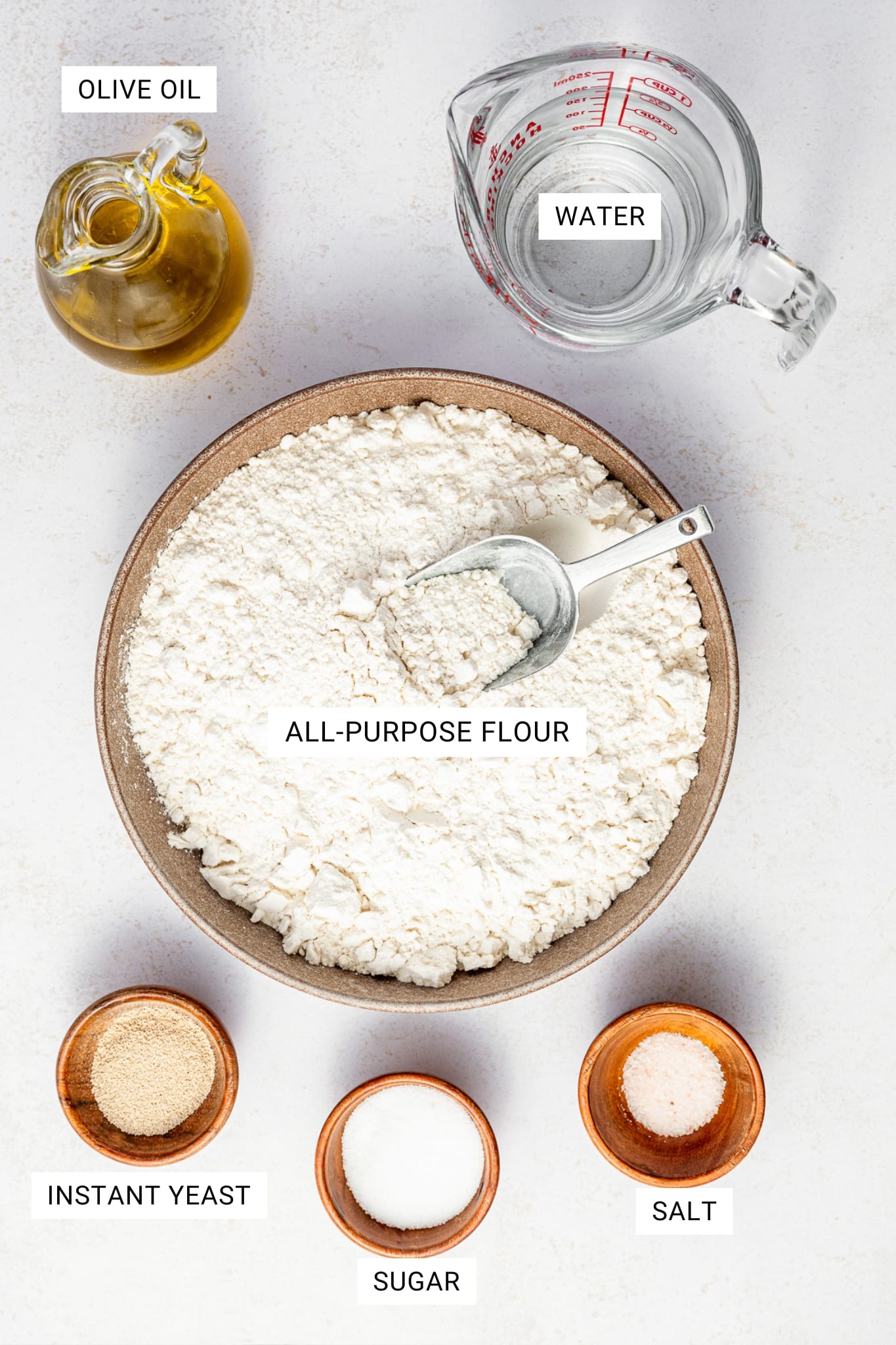 Overhead ingredient photo for homemade pita bread showing a tan bowl filled with all-purpose flour and a metal measuring spoon, a glass measuring cup of water, a glass cruet of olive oil, and three small wooden bowls holding instant yeast, sugar, and salt. White text overlays label each ingredient: "OLIVE OIL," "WATER," "ALL-PURPOSE FLOUR," "INSTANT YEAST," "SUGAR," and "SALT."