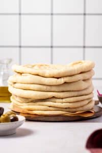 Tall stack of soft homemade pita bread on a round wooden board lined with brown parchment paper, with a white tile background, a glass bottle of olive oil on the left, a white bowl of green and Kalamata olives in the foreground, and a small bowl of pink pickled onions on the right.