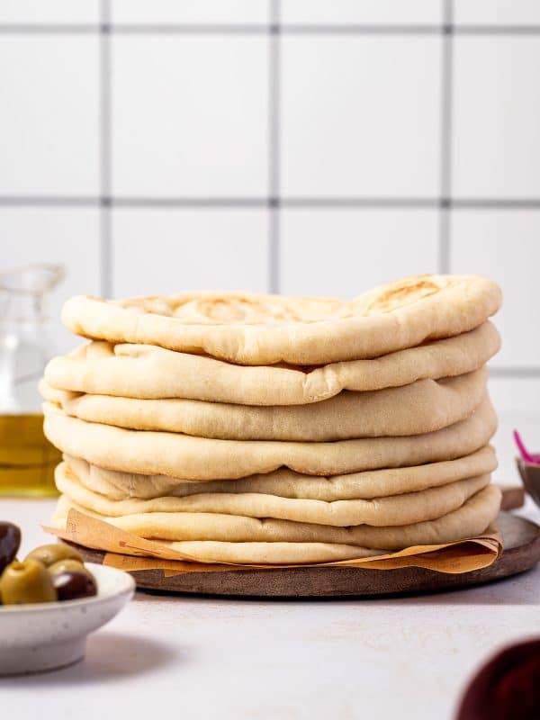 Tall stack of soft homemade pita bread on a round wooden board lined with brown parchment paper, with a white tile background, a glass bottle of olive oil on the left, a white bowl of green and Kalamata olives in the foreground, and a small bowl of pink pickled onions on the right.
