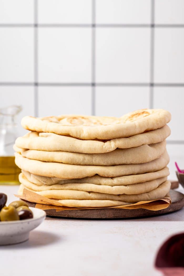Tall stack of soft homemade pita bread on a round wooden board lined with brown parchment paper, with a white tile background, a glass bottle of olive oil on the left, a white bowl of green and Kalamata olives in the foreground, and a small bowl of pink pickled onions on the right.