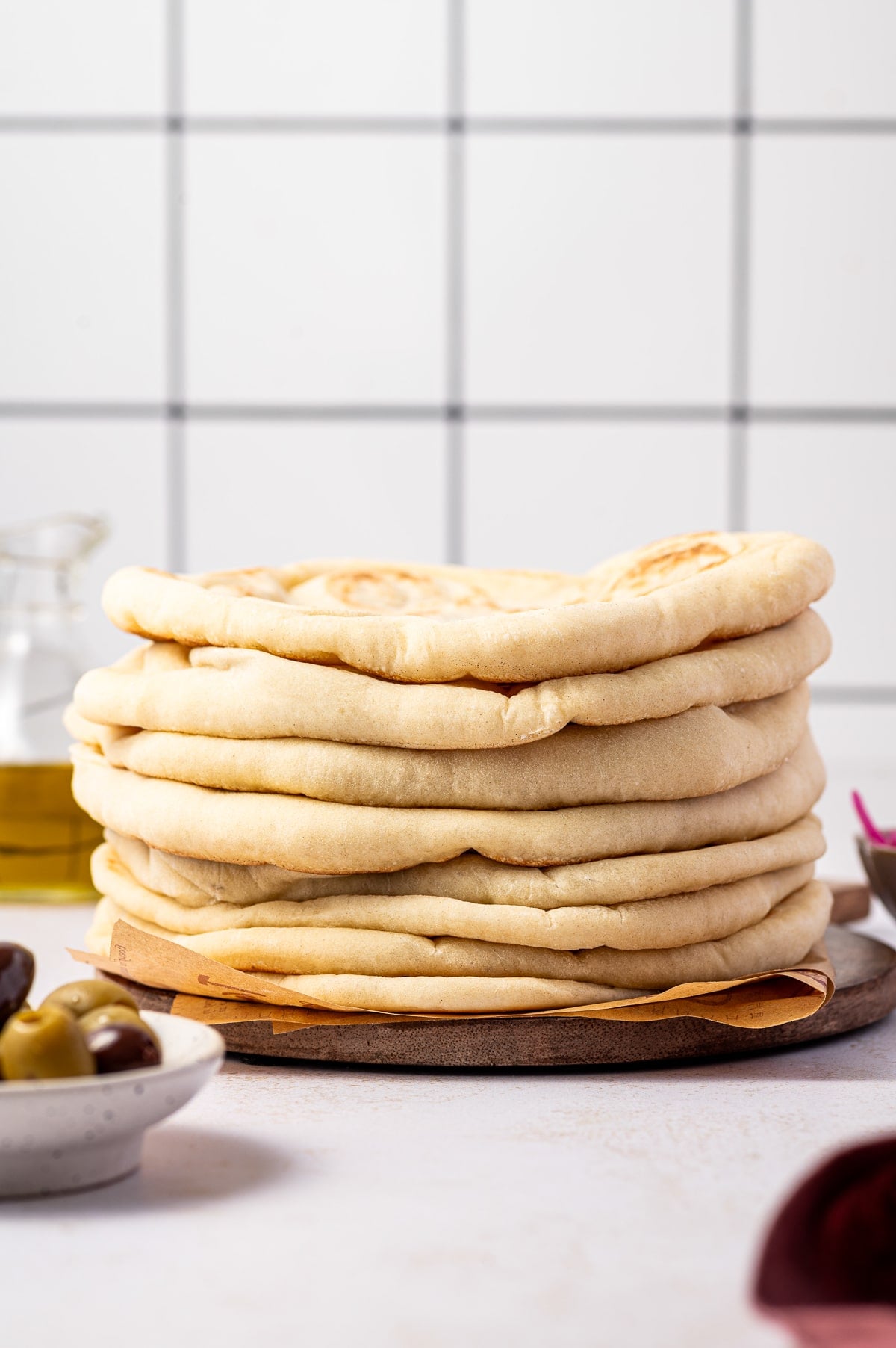 Tall stack of soft homemade pita bread on a round wooden board lined with brown parchment paper, with a white tile background, a glass bottle of olive oil on the left, a white bowl of green and Kalamata olives in the foreground, and a small bowl of pink pickled onions on the right.