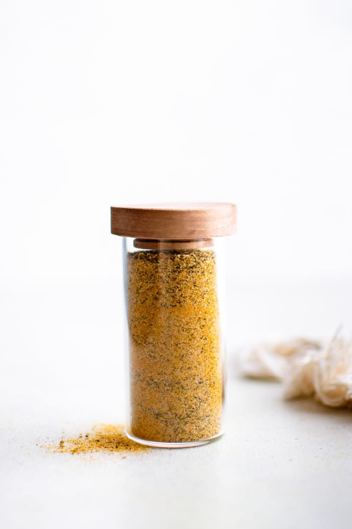 A tall glass spice jar with a wooden lid is filled to the top with homemade adobo all-purpose seasoning. A small pile of the spice blend sits beside the jar on a white surface.
