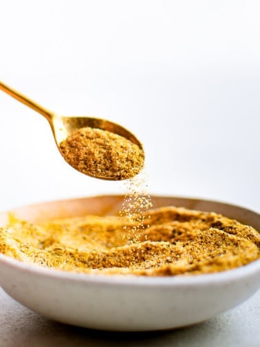 A gold spoon lifts a spoonful of homemade adobo seasoning from a white bowl as the spice gently cascades back down.