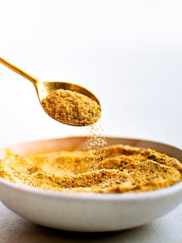 A gold spoon lifts a spoonful of homemade adobo seasoning from a white bowl as the spice gently cascades back down.