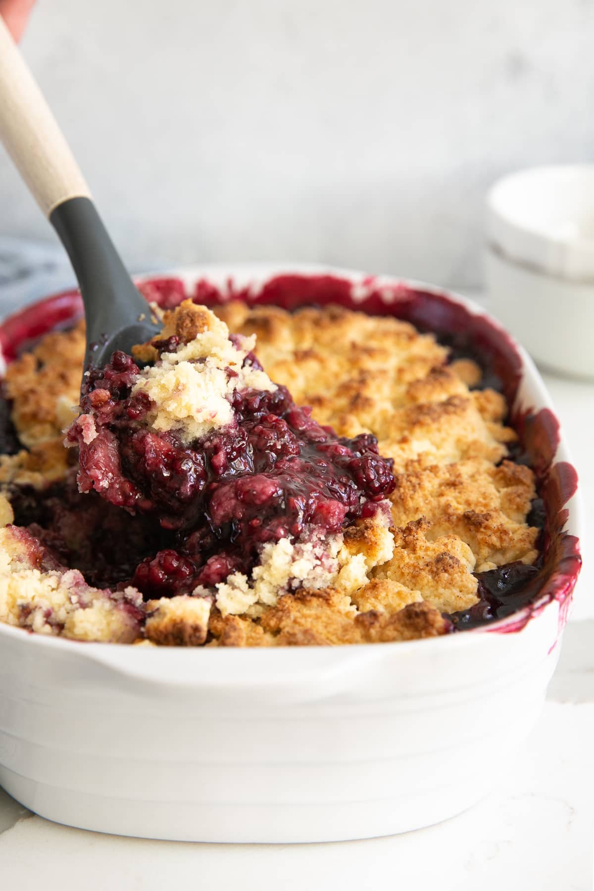 A bubbling blackberry cobbler with a golden, biscuit-style topping being scooped from a white casserole dish. The dark purple filling contrasts beautifully with the crispy top crust.