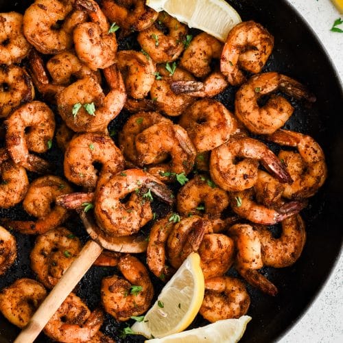 A rustic wooden spoon scoops up perfectly seared shrimp from a pan of blackened shrimp, with steam rising and lemon wedges on the side.