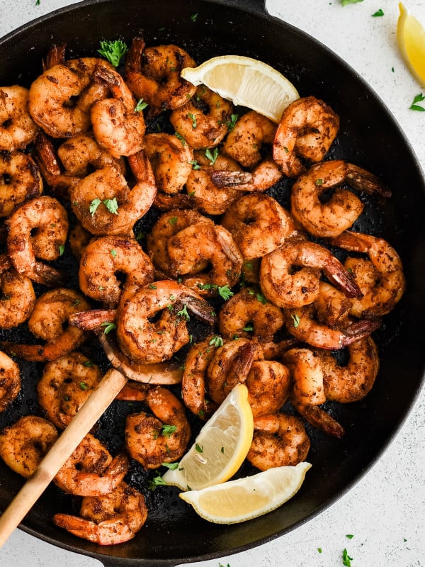 A rustic wooden spoon scoops up perfectly seared shrimp from a pan of blackened shrimp, with steam rising and lemon wedges on the side.
