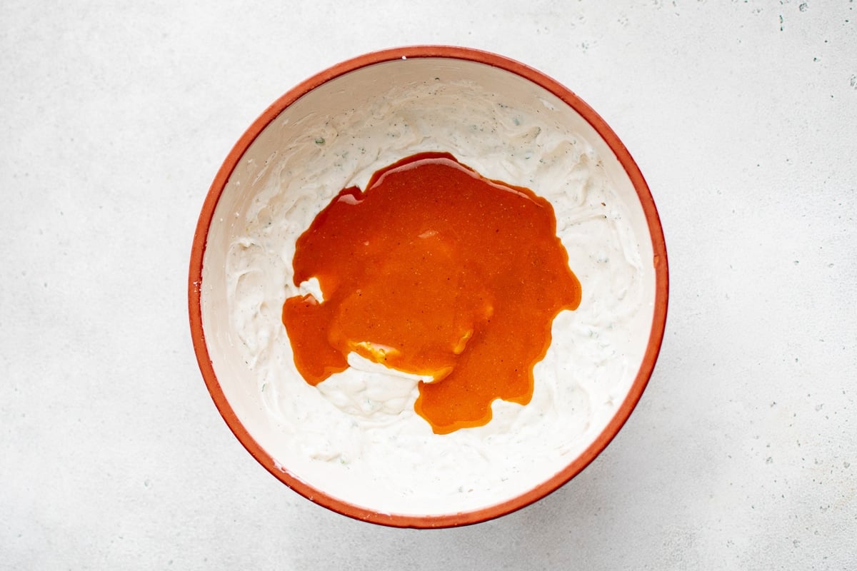 A bright orange buffalo sauce poured over a creamy ranch mixture in a mixing bowl.