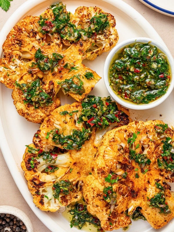 Overhead view of roasted cauliflower steaks covered in chimichurri sauce with a small bowl of extra sauce.