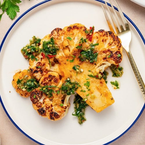 Single roasted cauliflower steak topped with chimichurri served on a round plate with a fork.