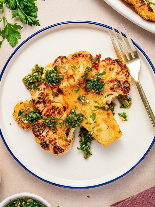 Single roasted cauliflower steak topped with chimichurri served on a round plate with a fork.