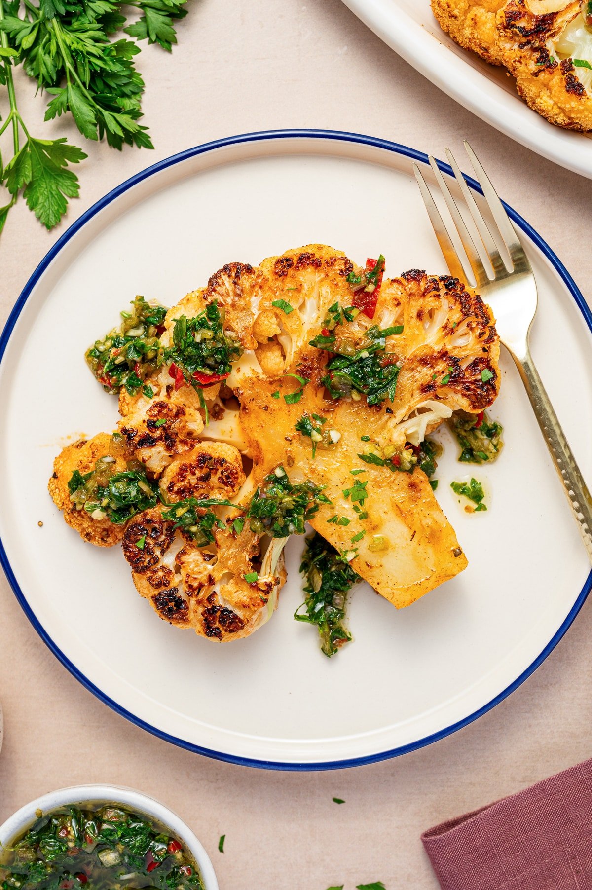 Single roasted cauliflower steak topped with chimichurri served on a round plate with a fork.