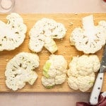 Sliced raw cauliflower steaks on a wooden cutting board next to a large chef’s knife.