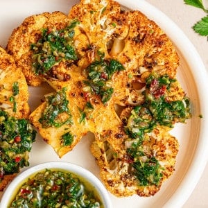 Square-cropped view of a single roasted cauliflower steak with chimichurri on a white plate with fork.