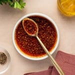 Small bowl of seasoned oil for cauliflower steaks, with a spoon resting inside and herbs visible throughout.