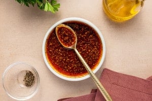 Small bowl of seasoned oil for cauliflower steaks, with a spoon resting inside and herbs visible throughout.