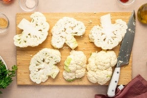 Sliced raw cauliflower steaks on a wooden cutting board next to a large chef’s knife.