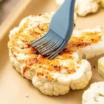 Cauliflower steak being brushed with seasoned oil using a silicone brush on a parchment-lined baking sheet.