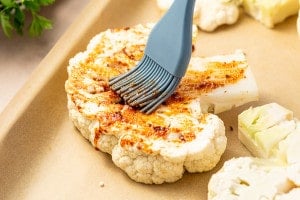 Cauliflower steak being brushed with seasoned oil using a silicone brush on a parchment-lined baking sheet.