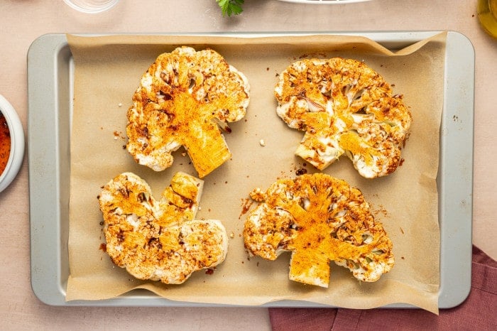 Four seasoned cauliflower steaks arranged on a parchment-lined baking sheet, ready to roast.