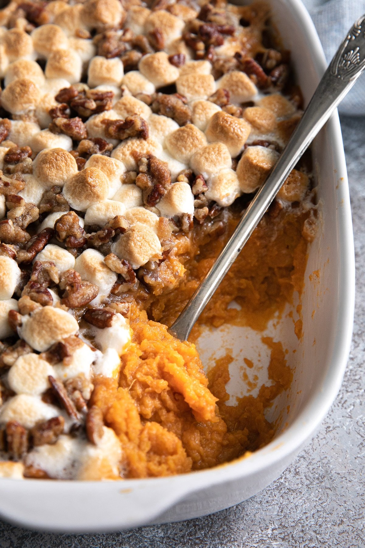 A rich and creamy sweet potato casserole topped with golden-brown marshmallows and a crunchy pecan streusel, with a serving spoon revealing the velvety filling beneath.