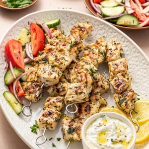 Angled view of a platter loaded with grilled chicken souvlaki skewers, lemon slices, tzatziki, and Greek salad with tomatoes, cucumbers, and red onions.